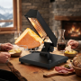 Appareil à raclette noir posé au centre d’une table en bois dans une ambiance montagnarde chaleureuse. Un quart de meule fond so