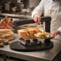 Dans une cuisine professionnelle, un cuisinier utilise un appareil à raclette noir pour faire fondre une demi-meule de fromage. 