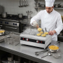 Un chef cuisinier en tenue blanche utilise un cuiseur à pâtes professionnel en inox dans une cuisine de restaurant. Il manipule 