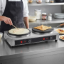 Crêpière professionnelle double en inox installée sur un plan de travail dans une cuisine. À gauche, un cuisinier étale de la pâ
